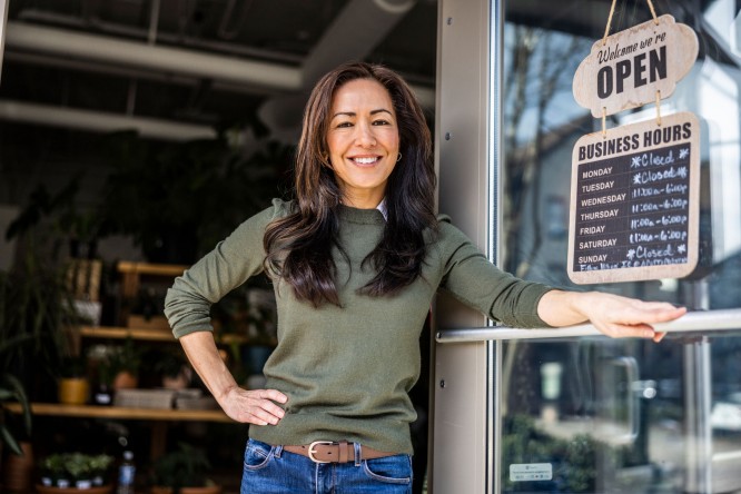 Frau hält Tür zu ihrem Geschäft offen, darauf hängt ein "Open"-Schild